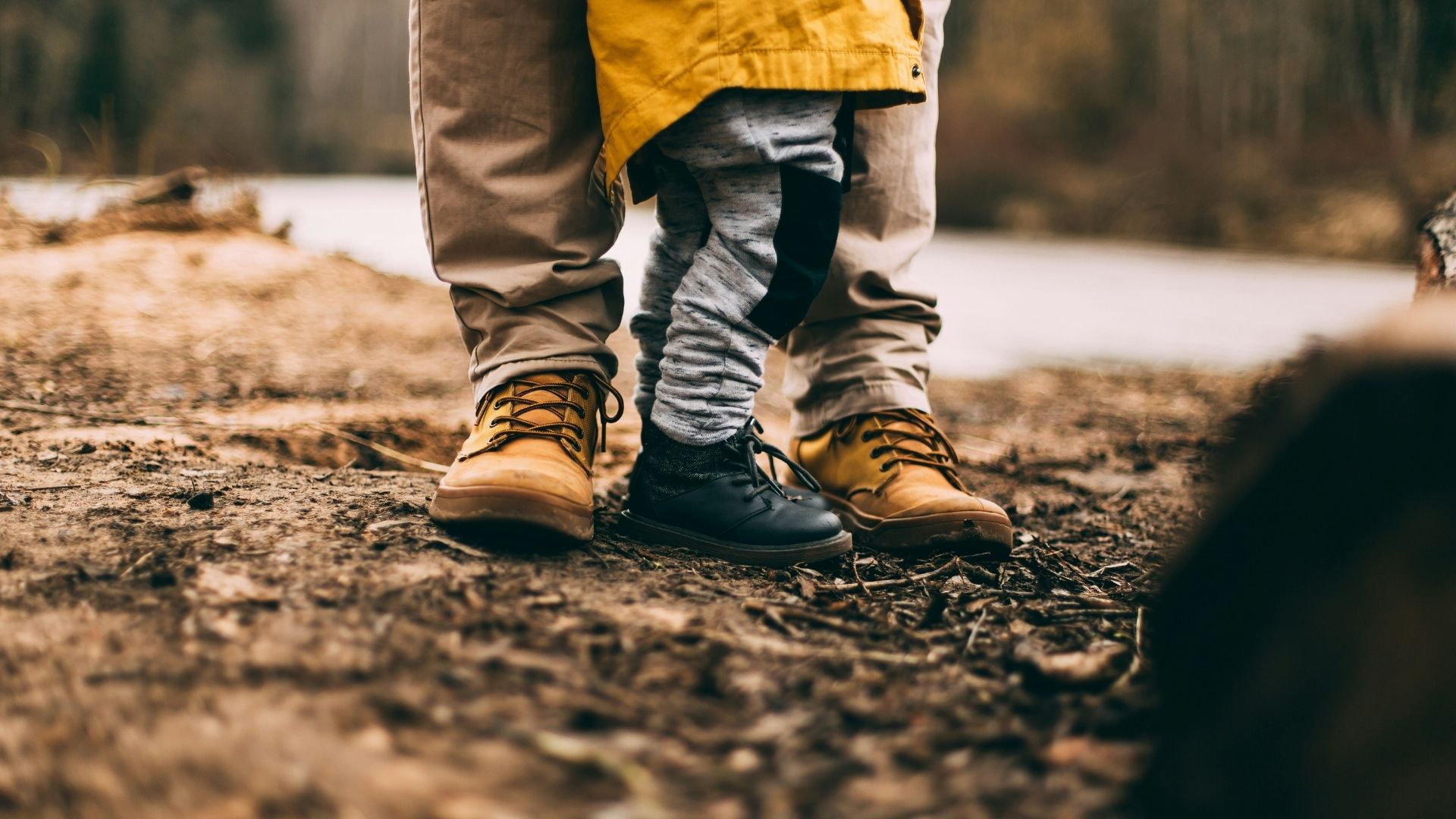 Das Bild zeigt die Füße eines Erwachsenen und eines Kindes, die nebeneinander stehen. Der Erwachsene trägt beige Hosen und braune Schnürstiefel, das Kind graue Hosen und schwarze Schnürschuhe. Der Hintergrund ist unscharf und zeigt eine natürliche Umgebung, möglicherweise eine Flusslandschaft. Der Fokus liegt auf den Schuhen und vermittelt ein Gefühl von Nähe und Verbundenheit.